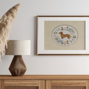 May include: Framed embroidery art featuring a brown dachshund dog surrounded by blue and pink flowers. The artwork is displayed in a brown frame with a white mat. The piece is set on a wooden cabinet, with a lamp and decorative plants.