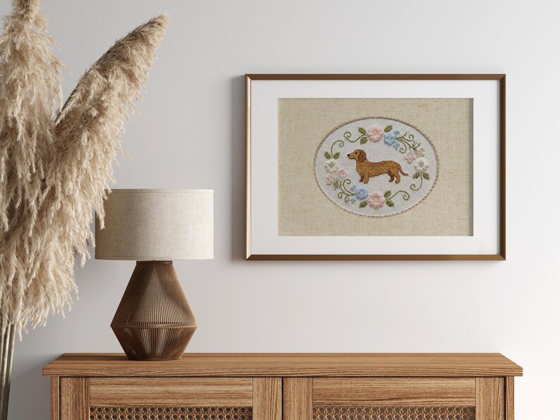 May include: Framed embroidery art featuring a brown dachshund dog surrounded by blue and pink flowers. The artwork is displayed in a brown frame with a white mat. The piece is set on a wooden cabinet, with a lamp and decorative plants.