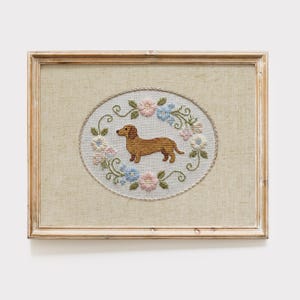 May include: Framed cross-stitch art featuring a brown dachshund dog inside an oval floral wreath. The wreath includes pink, blue, and white flowers with green leaves. The frame is a light brown color.
