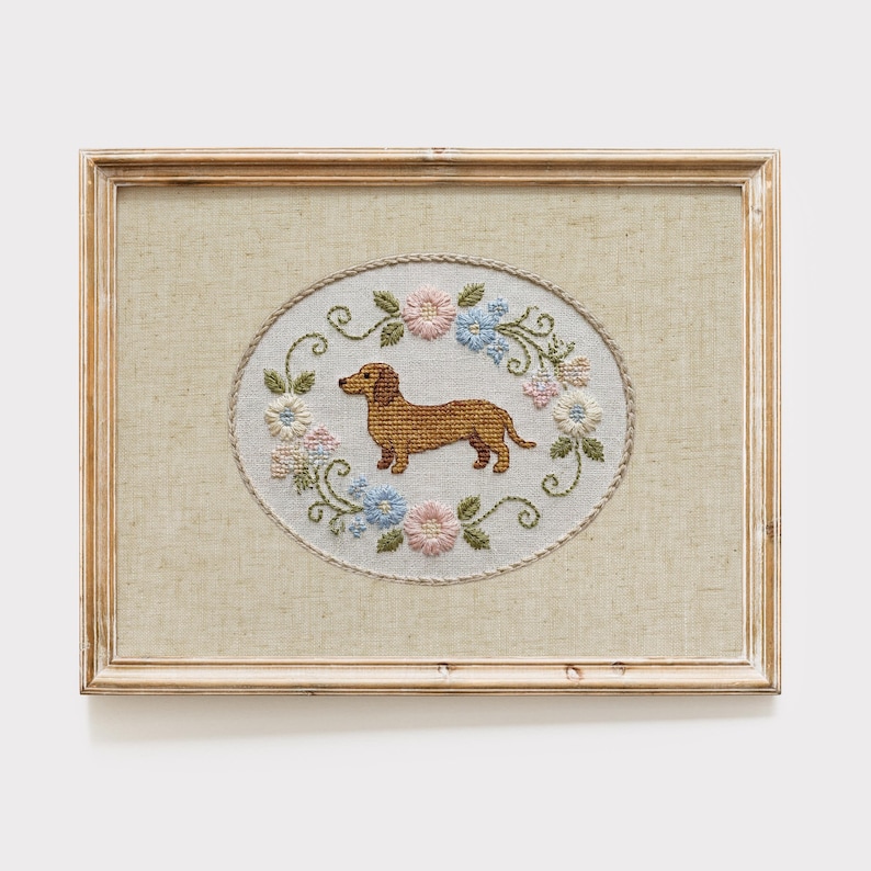 May include: Framed cross-stitch art featuring a brown dachshund dog inside an oval floral wreath. The wreath includes pink, blue, and white flowers with green leaves. The frame is a light brown color.