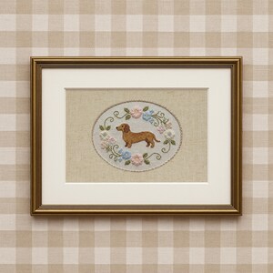 May include: Framed artwork featuring a cross-stitched dachshund dog surrounded by a floral wreath in pastel colors. The artwork is set against a neutral background and framed in a gold-colored frame. The overall aesthetic is vintage and charming.