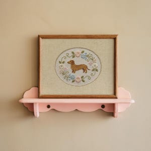 May include: A framed cross-stitch featuring a brown dachshund dog surrounded by a floral wreath in pastel colors. The artwork is displayed on a pink, scalloped-edge shelf, mounted on a beige wall. The frame is made of wood.