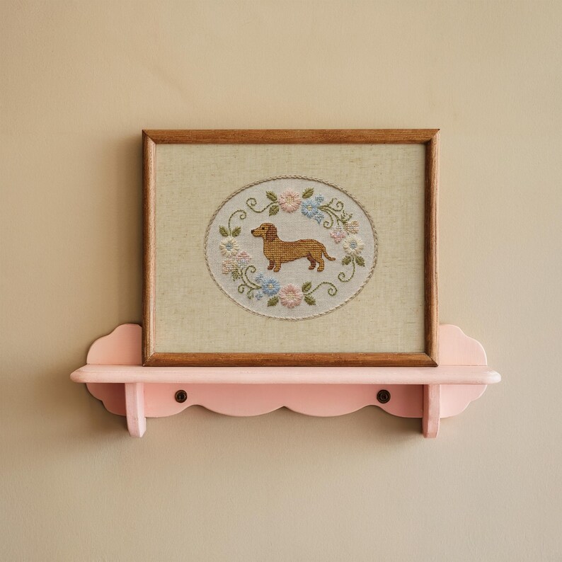 May include: A framed cross-stitch featuring a brown dachshund dog surrounded by a floral wreath in pastel colors. The artwork is displayed on a pink, scalloped-edge shelf, mounted on a beige wall. The frame is made of wood.