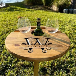 May include: A round, wooden wine picnic table with two stemless wine glasses and a wine bottle. The table has cutouts for the glasses and bottle, and a decorative design with the text "WOLF". The table is set on a grassy lawn.