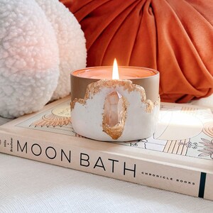 Op de afbeelding: Een witte kaars met een ingebedde heldere kwartskristal aan de zijkant. De kaars is aangestoken en heeft een warme, oranje gloed. De kaars staat op een boek met de titel "Moon Bath" met de woorden "Chronicle Books" gedrukt op de rug.