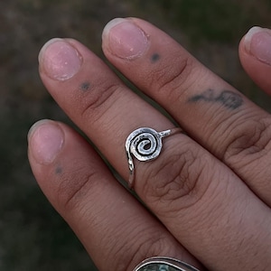 May include: A hand displays three silver rings. One ring features a spiral design, another has a sunburst pattern, and the third has a large oval green stone. The rings are set against a blurred outdoor background.