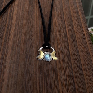 May include: A pendant necklace featuring a central round iridescent moonstone cabochon flanked by two gold-toned crescent moon shapes, all set within a silver-toned frame, suspended from a simple black cord.