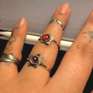 May include: Two silver rings with star-shaped settings. One ring features a red gemstone, the other a purple gemstone. Additional silver rings are also visible on the fingers.