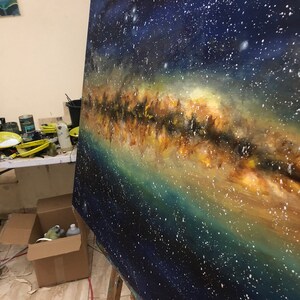 May include: A close-up of an oil painting depicting the Milky Way galaxy. The painting features a dark blue background with a band of white, yellow, and orange stars. The painting is on an easel in a studio setting.