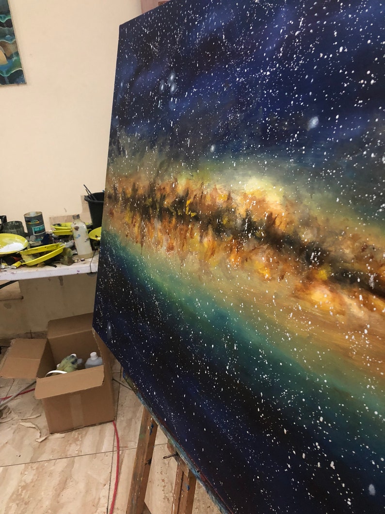 May include: A close-up of an oil painting depicting the Milky Way galaxy. The painting features a dark blue background with a band of white, yellow, and orange stars. The painting is on an easel in a studio setting.