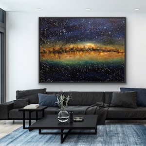 May include: A large abstract painting of a starry night sky with a band of golden and green light across the center. The painting is framed in black and hanging on a white wall in a modern living room.