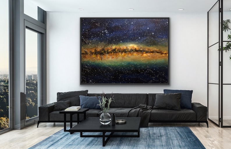May include: A large abstract painting of a starry night sky with a band of golden and green light across the center. The painting is framed in black and hanging on a white wall in a modern living room.