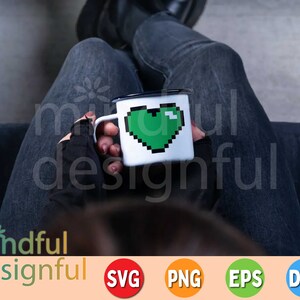 May include: A white enamel mug with a black rim and handle, featuring a pixelated green heart design. The mug is held by a person wearing black gloves and dark jeans. The image includes the text "mindful designful".