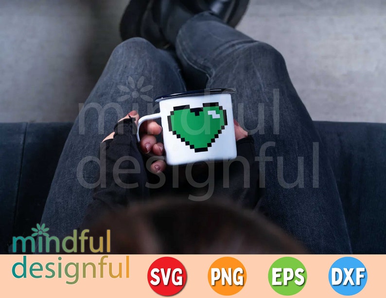 May include: A white enamel mug with a black rim and handle, featuring a pixelated green heart design. The mug is held by a person wearing black gloves and dark jeans. The image includes the text "mindful designful".