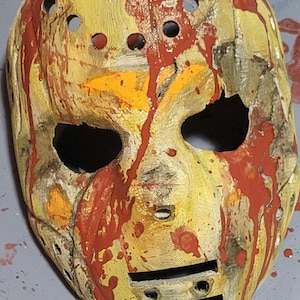 May include: A hockey mask painted with red and orange paint splatters. The mask has a distressed look with visible cracks and holes.