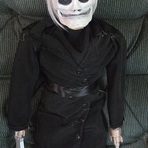 May include: A doll of a character from a horror film, wearing a black hat and coat. The doll has a white skull-like face, white hair, and silver-colored hands holding a knife and a hook.