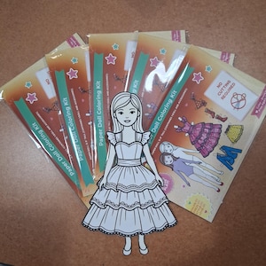 May include: A paper doll coloring kit featuring a die-cut paper doll in a tiered dress placed over several identical packaged kits. Each package displays the text Paper Doll Coloring Kit, 8 inch tall, and No Cutting Required with star graphics.