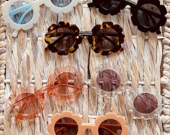 May include: A set of seven pairs of flower-shaped sunglasses in various colors, including white, black, brown, pink, and clear. The sunglasses are arranged on a woven wicker surface.
