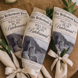 Könnte beinhalten: Drei zeitungartige Karten mit dem Titel The Bridesmaid Post und dem Text Will You Be My Bridesmaid, die ein schwarz-weißes Foto von zwei Personen zeigen, eingewickelt in cremefarbenes Papier mit passendem Band und grünen Blattakzenten.