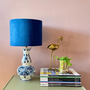 Può includere: Un paralume in velluto blu è posto su una base di lampada in ceramica floreale bianca e blu. Una statua di fenicottero dorato e una piccola pianta in vaso sono su riviste su una cassettiera verde.