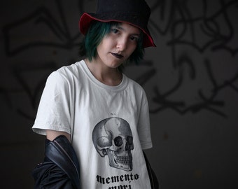 May include: A person wearing a black bucket hat with a red brim, a white t-shirt with a black and white skull graphic and the text "memento mori", and a black leather jacket.