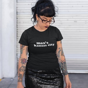 May include: A black t-shirt with the text "max's kansas city" in white lettering. The person wearing the shirt has black hair, black framed glasses, and tattoos on their arms.