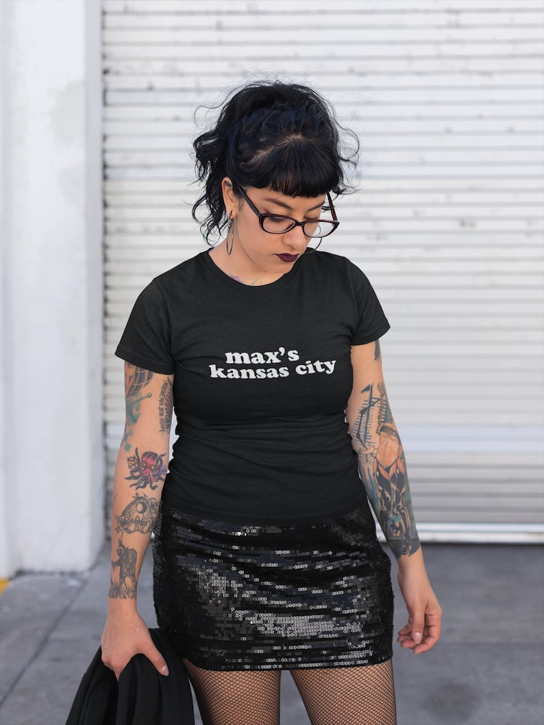 May include: A black t-shirt with the text "max's kansas city" in white lettering. The person wearing the shirt has black hair, black framed glasses, and tattoos on their arms.