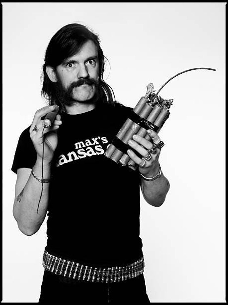 May include: A black and white photo of a person wearing a black t-shirt with the text "max's ansas" printed on it. The person is holding a bundle of dynamite with a lit fuse.