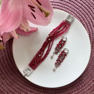 May include: A set of red gemstone jewelry featuring a multi-strand bracelet and dangling earrings. The bracelet has a silver clasp and the earrings have silver hooks.