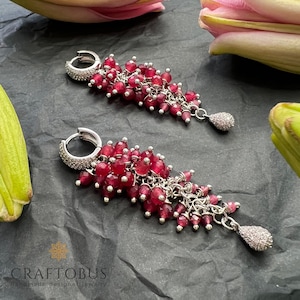 May include: A pair of silver hoop earrings with a dangling cluster of red gemstone beads and a silver teardrop charm. The earrings are on a dark gray surface.
