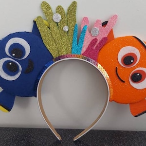 May include: A headband with a blue Dory and an orange Nemo character from the movie Finding Nemo. The headband is decorated with a sparkly gold and pink coral.