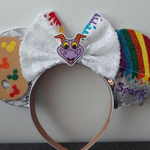 May include: A pair of white sparkly mouse ears with a purple dragon character, a paint palette, and a rainbow paint splatter design. The ears are decorated with colorful paint splatters and the text "Spark".