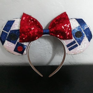 May include: A pair of white and blue mouse ears with a red bow. The ears feature a white background with blue and black geometric shapes. The bow is made of red sequins.