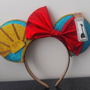 May include: A pair of turquoise blue mouse ears with gold glitter trim and a red bow. One ear features a gold sunburst design and the other ear has a white bottle with a black giraffe silhouette.