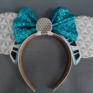 May include: A pair of white and gray mouse ears with a turquoise bow and a silver Spaceship Earth charm. The ears are decorated with a geometric pattern.