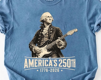 Puede incluir: Camiseta azul con una ilustración de George Washington tocando una guitarra eléctrica. El diseño incluye el texto "AMERICA'S 250TH" y los años "1776-2026". La camiseta está hecha de un material suave.