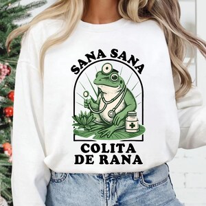 May include: White sweatshirt featuring a cartoon frog doctor with a stethoscope, holding a magnifying glass, and a medicine bottle. The design includes the text "SANA SANA" above and "COLITA DE RANA" below the frog illustration.