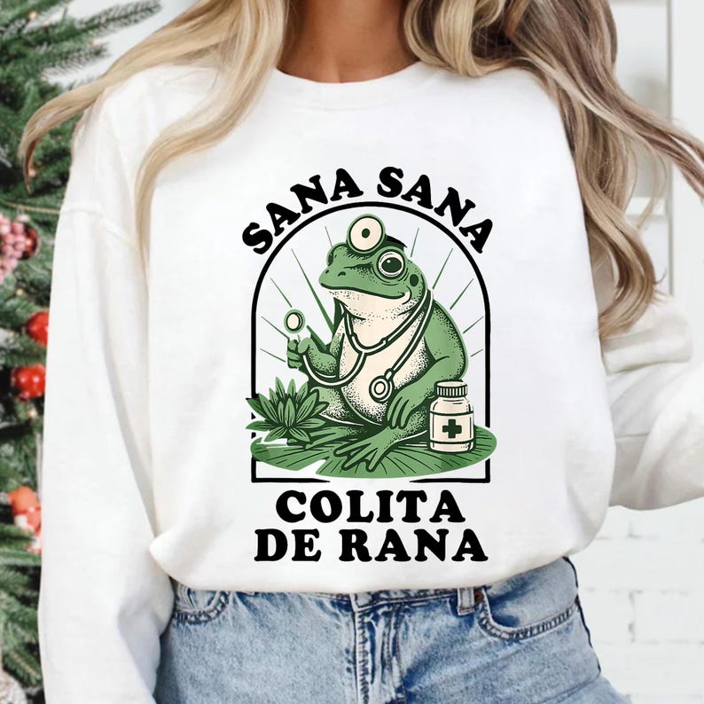 May include: White sweatshirt featuring a cartoon frog doctor with a stethoscope, holding a magnifying glass, and a medicine bottle. The design includes the text "SANA SANA" above and "COLITA DE RANA" below the frog illustration.