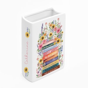 May include: White ceramic book-shaped vase with a floral design and the name "Catherine" in pink script. The floral arrangement includes sunflowers, roses, and other flowers. The vase features book titles like "Pride and Prejudice."