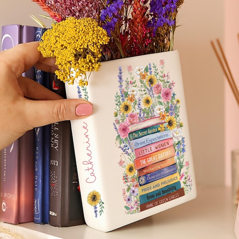 May include: A white ceramic vase shaped like a stack of books, adorned with floral illustrations and book titles. The vase is personalized with the name "Catherine" and filled with colorful dried flowers. The titles include "The Secret Garden" and "Pride and Prejudice".