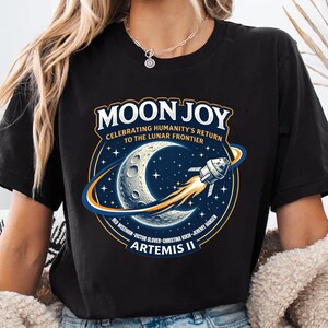 May include: Black t-shirt featuring a circular graphic with the words "MOON JOY" and "ARTEMIS II". The graphic includes a crescent moon, stars, and a rocket ship. The text "CELEBRATING HUMANITY'S RETURN TO THE LUNAR FRONTIER" is also present.