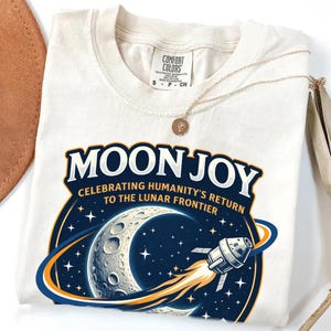 May include: Cream-colored t-shirt with a graphic design featuring the words "MOON JOY" above a crescent moon, stars, and a spaceship. The text "CELEBRATING HUMANITY'S RETURN TO THE LUNAR FRONTIER" is also included. A gold necklace is draped across the shirt.