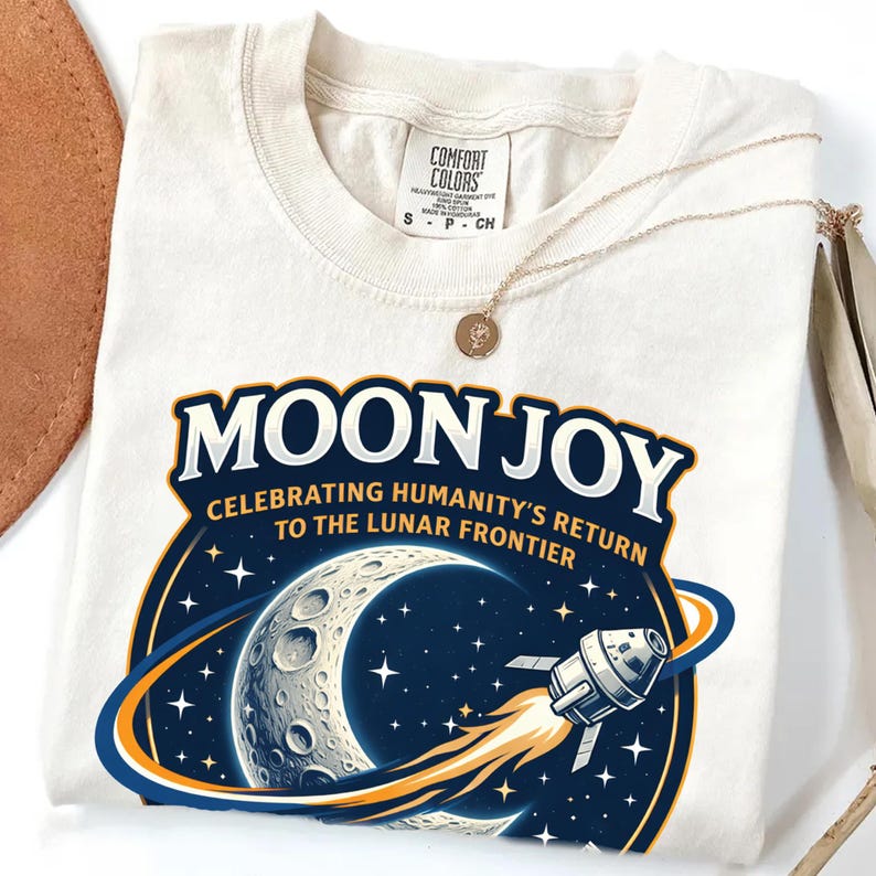 May include: Cream-colored t-shirt with a graphic design featuring the words "MOON JOY" above a crescent moon, stars, and a spaceship. The text "CELEBRATING HUMANITY'S RETURN TO THE LUNAR FRONTIER" is also included. A gold necklace is draped across the shirt.