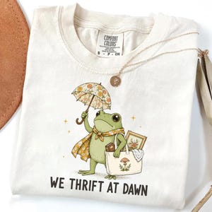 May include: A cream-colored short-sleeve t-shirt featuring a graphic of a green frog holding a floral umbrella and a tote bag containing a book and a framed picture. The text WE THRIFT AT DAWN is printed below the frog in a bold, dark font.