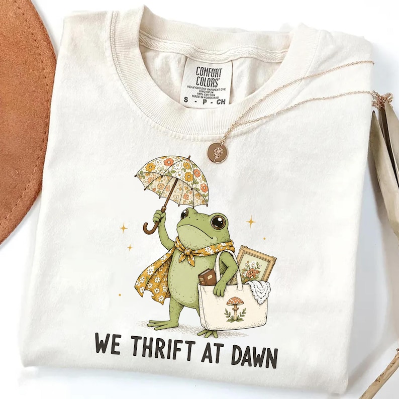 May include: A cream-colored short-sleeve t-shirt featuring a graphic of a green frog holding a floral umbrella and a tote bag containing a book and a framed picture. The text WE THRIFT AT DAWN is printed below the frog in a bold, dark font.