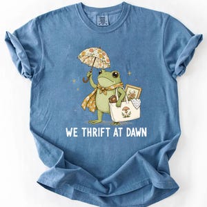 May include: A blue t-shirt featuring a graphic of a green frog holding a floral umbrella and carrying a white tote bag with a mushroom illustration. The frog wears a floral scarf. Below the graphic, the text reads WE THRIFT AT DAWN.
