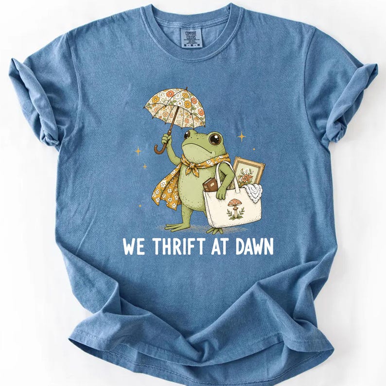 May include: A blue t-shirt featuring a graphic of a green frog holding a floral umbrella and carrying a white tote bag with a mushroom illustration. The frog wears a floral scarf. Below the graphic, the text reads WE THRIFT AT DAWN.
