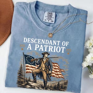 May include: Light blue Comfort Colors t-shirt featuring the text "Descendant of a Patriot" above an illustration of a colonial soldier holding a rifle and standing in front of a flag. A gold necklace with a pendant rests on the shirt.