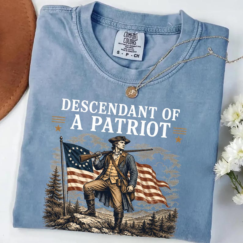 May include: Light blue Comfort Colors t-shirt featuring the text "Descendant of a Patriot" above an illustration of a colonial soldier holding a rifle and standing in front of a flag. A gold necklace with a pendant rests on the shirt.