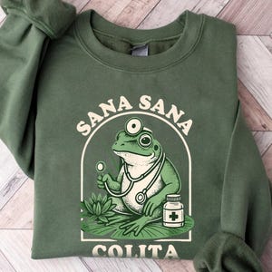 May include: Green sweatshirt featuring a cartoon frog doctor with a stethoscope and a pill bottle. The frog is inside a white arched frame with the words "Sana Sana Colita" in white text. The sweatshirt is a solid green color.